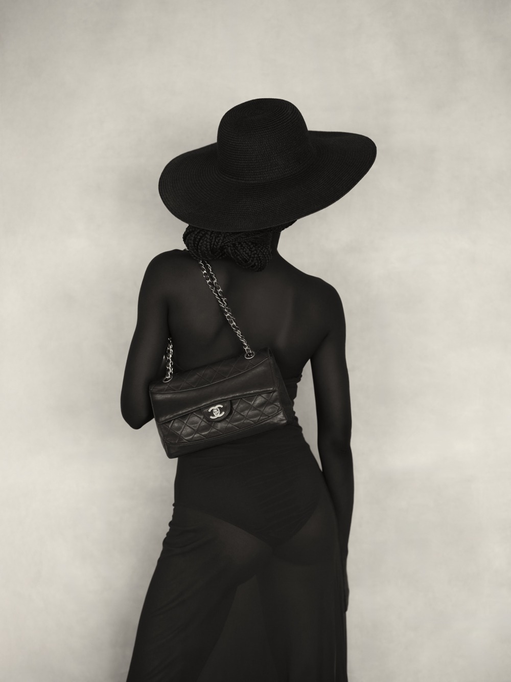 A person seen from behind, dressed in a dark sheer dress and black hat, holding a small chanel quilted handbag with a chain strap.