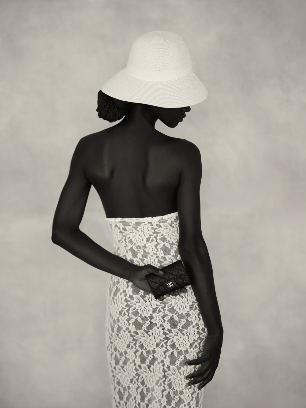 A person wearing a strapless, white lace dress and a white wide-brimmed hat, standing against a light neutral backdrop, holding a chanel bag.