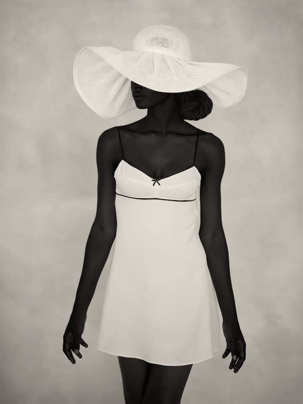 A person in a white sleeveless dress and a large floppy hat with a bow, standing against a pale background.