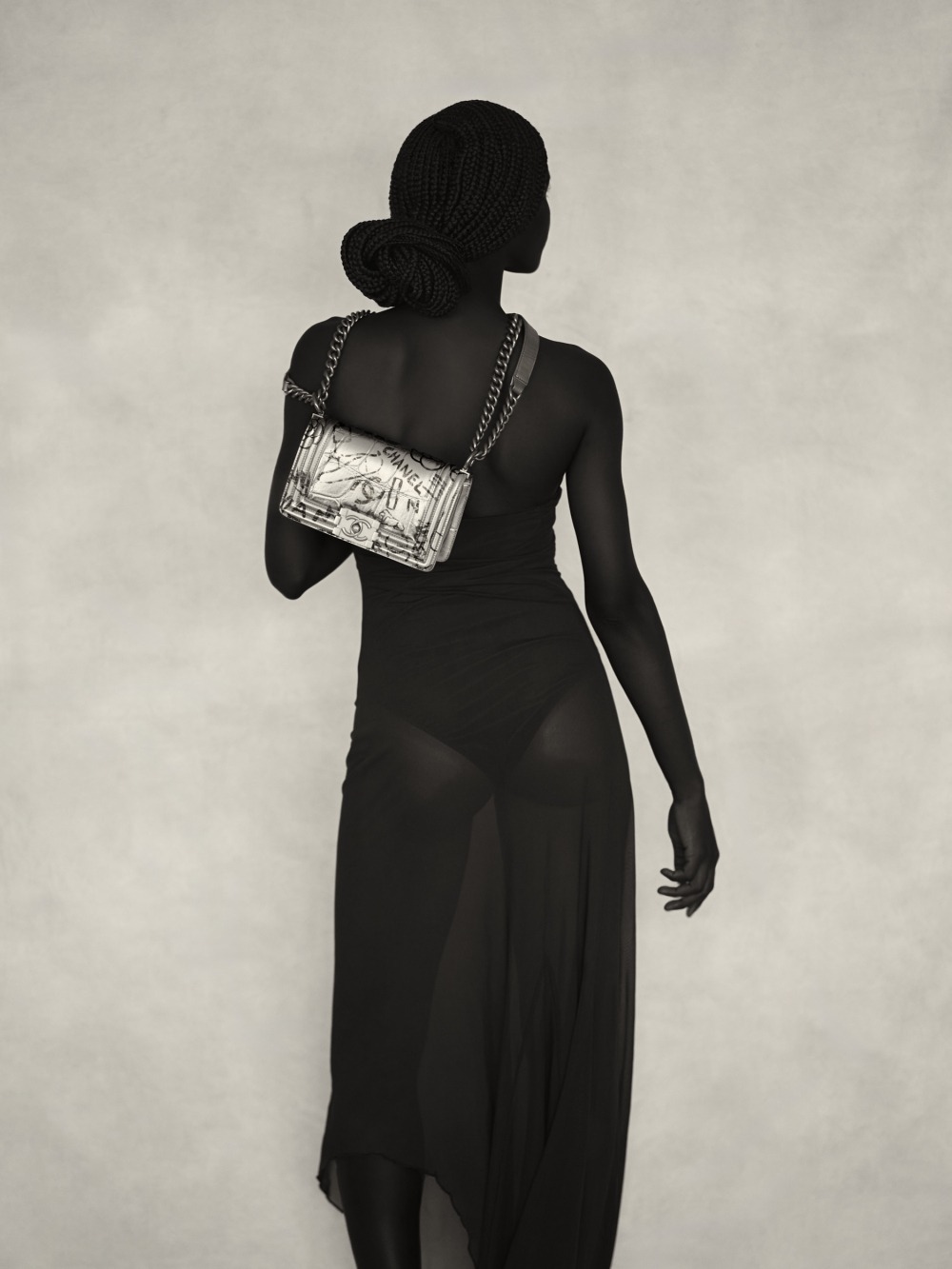 A woman seen from behind, wearing a black sheer dress and carrying a silver metallic chanel handbag, with their head slightly turned.