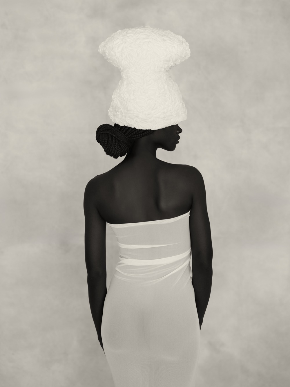 A person with dark skin, draped in a white fabric and wearing a textured white headwrap, standing against a plain background.
